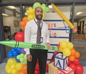 Picture of a man holding a balloon crayon standing in front of a back to school photo back drop