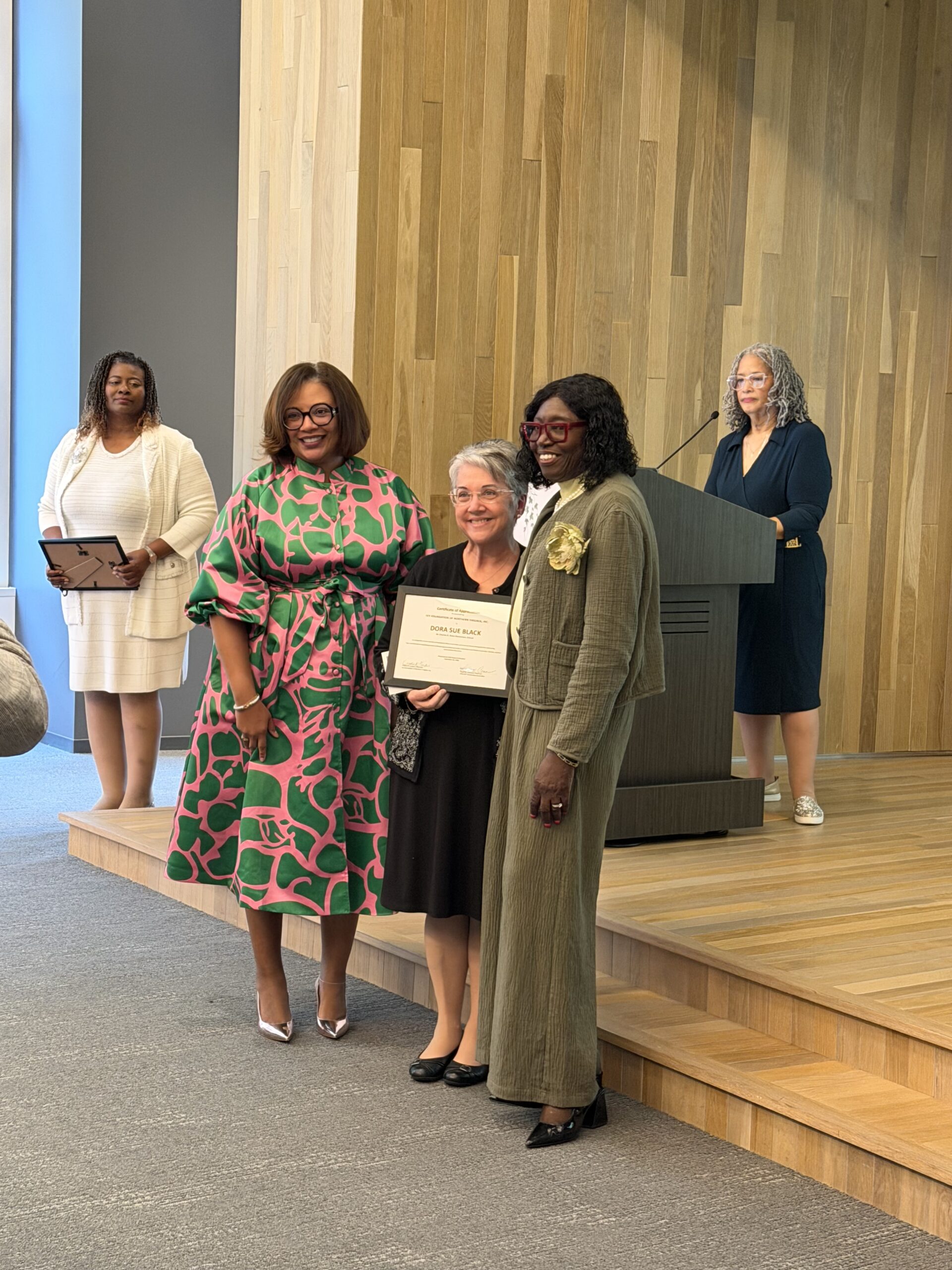 A picture of 5 women standing on a stage. One woman is holding a certificate as an award to honor her service in the library.